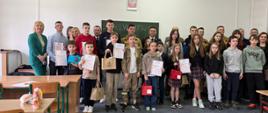 Dzieci trzymające dyplomy i nagrody za nimi stoją dorośli ludzie. W tle widać zieloną tablicę szkolną. Nad nią wisi Godło Polski.