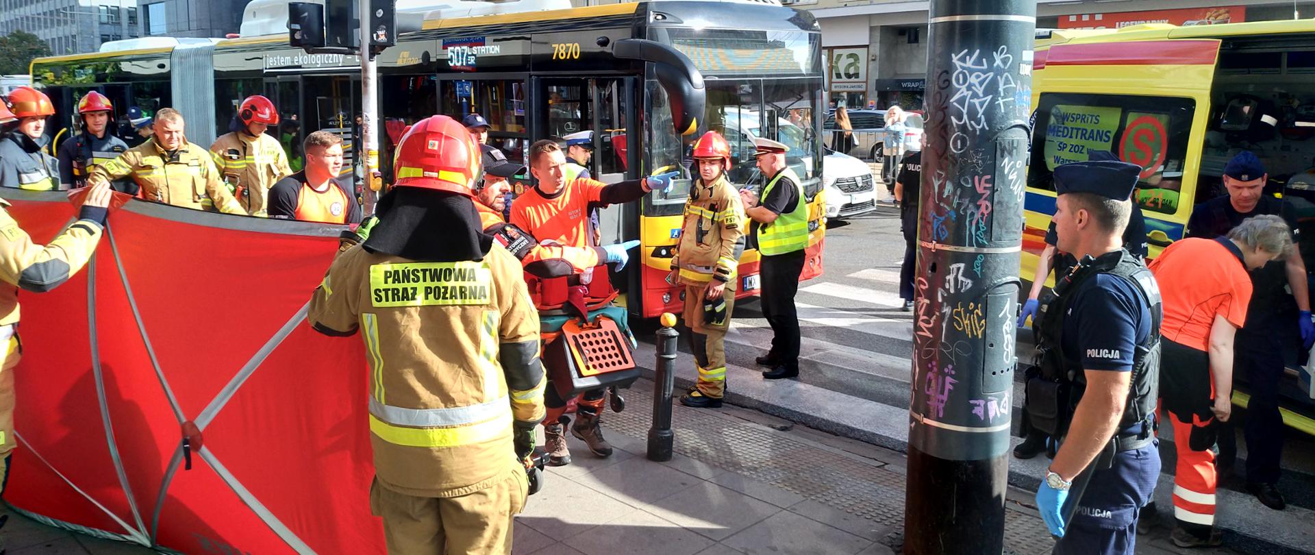 Widoczni strażacy trzymający parawan, ratownicy medyczni w trakcie przenoszenia poszkodowanego na noszach do karetki, oraz policja zabezpieczająca miejsce zdarzenia.