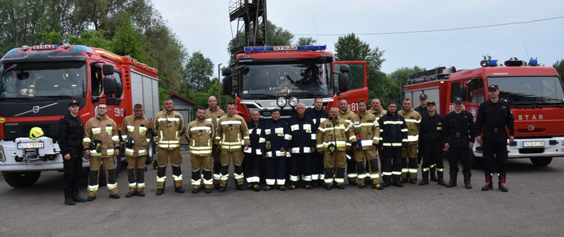 Pełne skład plutonu ratowniczego OSP wraz z funkcjonariuszami KP PSP Ostróda stoją przed samochodami ratowniczo-gaśniczymi. 