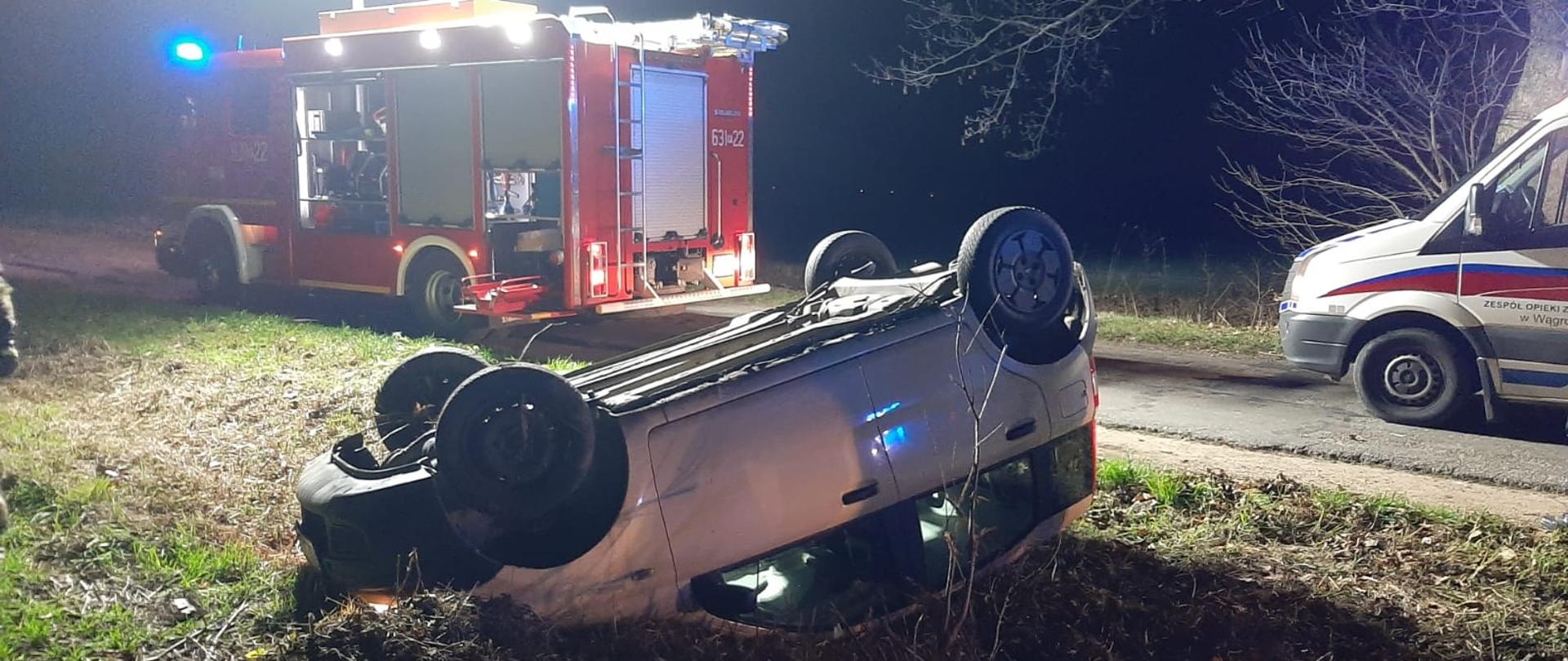 Na zdjęciu widać samochód przewrócony na dach oraz wóz straży pożarnej i karetkę
