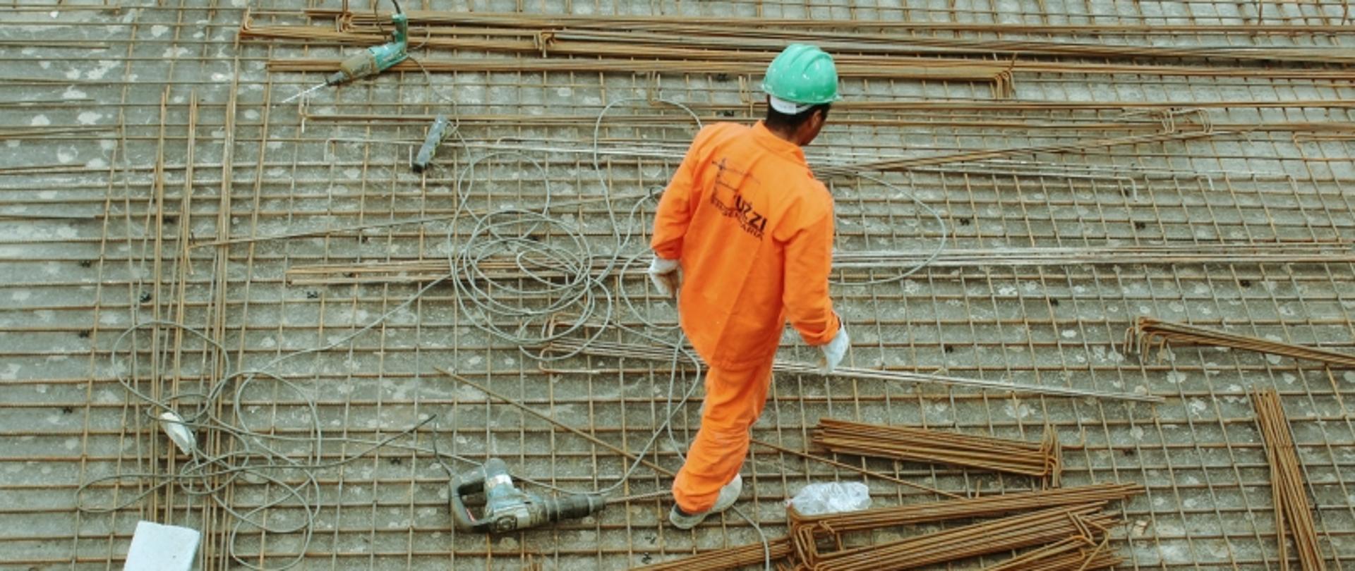 Robotnik na placu budowy stojący na zbrojeniu, wokół metalowe pręty i sprzęt.