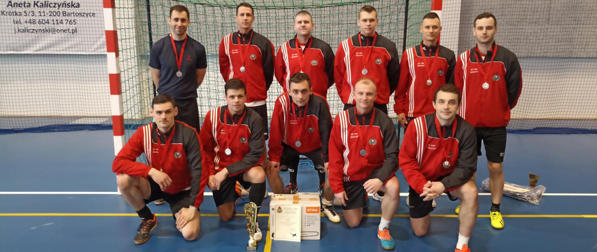 Zdjęcie przedstawia drużynę Komendy Powiatowej Państwowej Straży Pożarnej w Gołdapi (jedenastu zawodników) - Wicemistrzów Województwa Warmińsko–Mazurskiego w Halowej Piłce Nożnej ze zdobytymi trofeami, na tle bramki w hali widowiskowo–sportowej Bartoszyckiego Ośrodka Sportu i Rekreacji