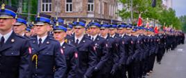 Kolumna funkcjonariuszy PSP w galowych mundurach przemieszcza się ulicą w zwartym szyku marszowym. Uczestnicy mają na sobie granatowe mundury z niebieskimi otokami czapek oraz godłem państwowym na rękawie. W tle widoczne są kamienice i witryny miejskie, co nadaje zdjęciu charakter podniosłego przemarszu przez centrum Warszawy.