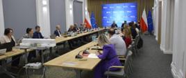 Zdjęcie od tyłu pod kątem, na sali za ustawionymi w czworokąt stołami siedzi dużo ludzi, u szczytu stołu wiceminister Szeptycki, za nim na ścianie wielki ekran, po obu jego stronach flagi Polski i UE.