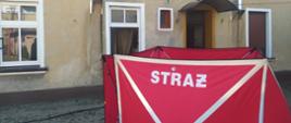 Tragiczny bilans pożaru. Posesja w Rawiczu przy ul. Piłsudskiego. Na podwórzu, przed kamienicą ustawiony jest parawan ochronny koloru czerwonego z białym napisem STRAŻ. Drzwi wejściowe oraz jedno z okien kamienicy są otwarte. 