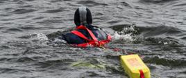 Strażak ubrany w piankę płynie do osoby poszkodowanej znajdującej się w wodzie. Strażak posiada czerwoną uprząż zabezpieczającą oraz pas ratunkowy "węgorz" koloru żółtego z czerwonym napisem "RESCUE" 