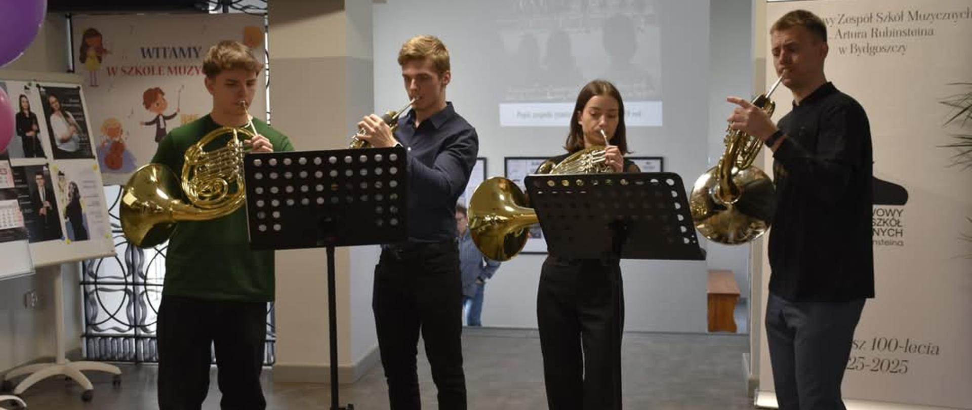 na zdjęciu trzech meżczyzn w czarnych koszulach i jedna kobieta grajacy na tubach w złotym kolorze nad nimi ekran w tle plkaty