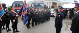 Na pierwszym planie, po lewej stronie, widoczny jest poczet sztandarowy . Obok niego w dwóch rzędach, stoi grupa strażaków w mundurach galowych, ustawionych w szyku. Po prawej stronie, naprzeciw nich, stoi strażak oddający honory. W tle widać dwa czerwone wozy strażackie oraz grupę gości, częściowo stojących pod namiotem. Jest widno.