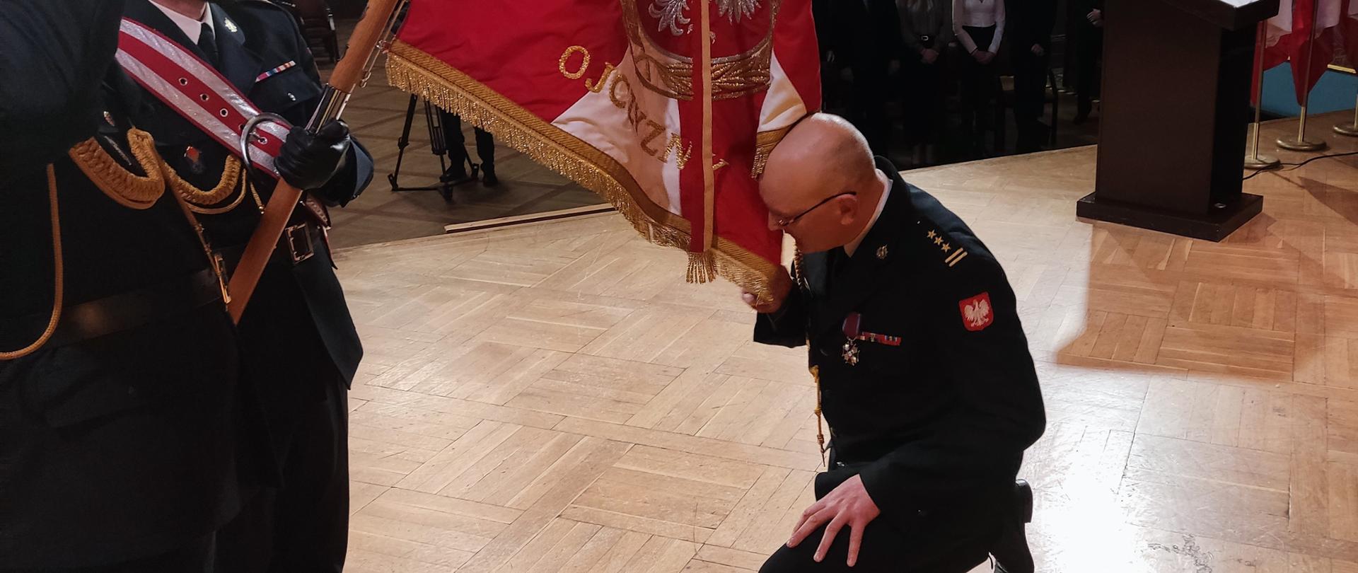 Komendant klęcząc całuje płat sztandaru. Z lewej strony widok na sztandarowego pochylającego sztandar. W tle widać gości uroczystości.