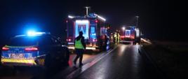 Dwa wozy strażackie oraz radiowóz policji podczas zdarzenia. Wozy strażackie oświetlają teren akcji za pomocą najaśnic.