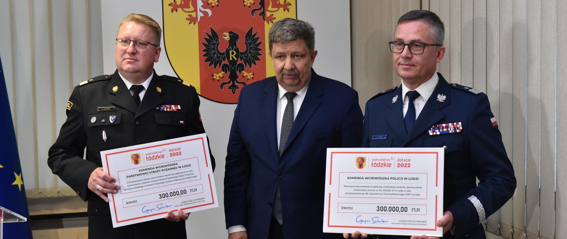 dwóch generałów straży pożarnej i policji oraz mężczyzna w garniturze, mężczyzna stoi pomiędzy generałami, którzy trzymają tabliczkę z kwotą dotacji 300000 złotych, w tle na ścianie herb województwa łódzkiego 