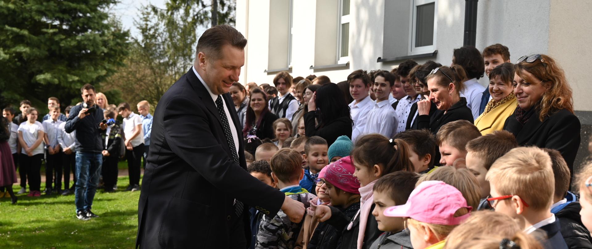 Minister przybija piątkę z małą dziewczynką, stojącą wśród dzieci pod ścianą budynku.