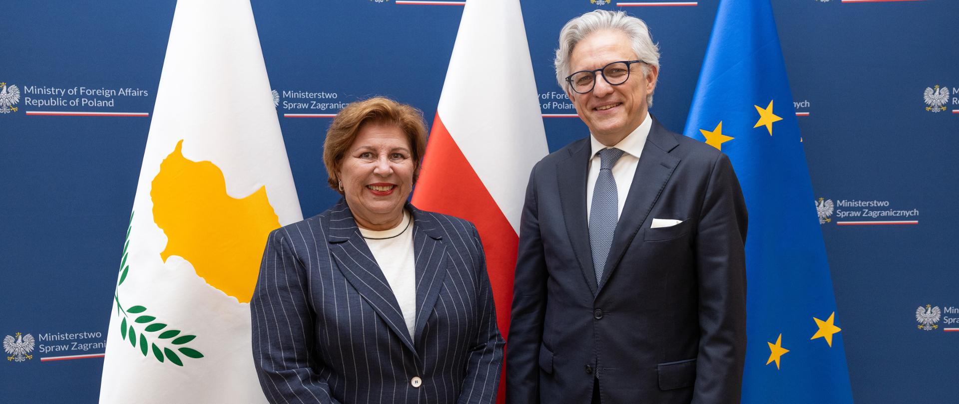 Podsekretarz Stanu Artur Harazim ze Stałą Sekretarz Ministerstwa Spraw Zagranicznych Republiki Cypryjskiej Theodorą Constantinidou uśmiechają się, pozując razem na tle flag oraz granatowej ściance MSZ.