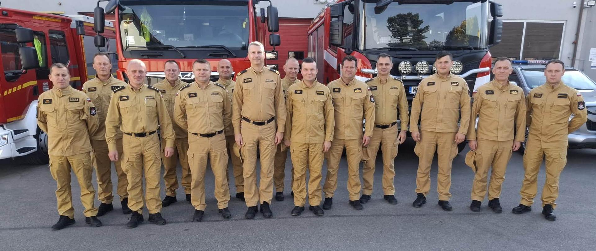 Pozowane zdjęcie grupowe trzynastu strażaków w piaskowych mundurach służbowych, stojących w jednym rzędzie przed placem manewrowym komendy. W tle zaparkowane są czerwone wozy strażackie Państwowej Straży Pożarnej oraz budynek komendy.
