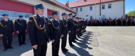 Zdjęcie przedstawia teren Jednostki Ratowniczo-Gaśniczej oraz strażaków ubranych w mundury galowe koloru czarnego ze sznurem galowym oraz czapką rogatywką na głowie, podczas obchodów Dnia Strażaka w Radziejowie. Na zdjęciu w pierwszym szeregu strażacy awansowani na wyższe stopnie służbowe
