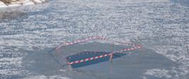 Na pierwszym planie zabezpieczona taśmą ostrzegawczą przerębla po skończonych ćwiczeniach strażaków. W tle aura śnieżna, staw skuty lodem.