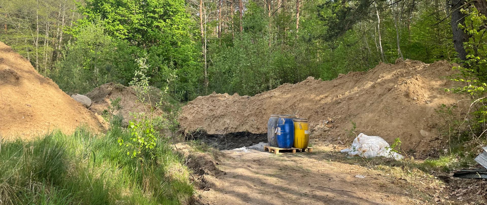 Widok na przygotowane do opróżnienia beczki oraz dół otoczony hałdami piasku i gleby.