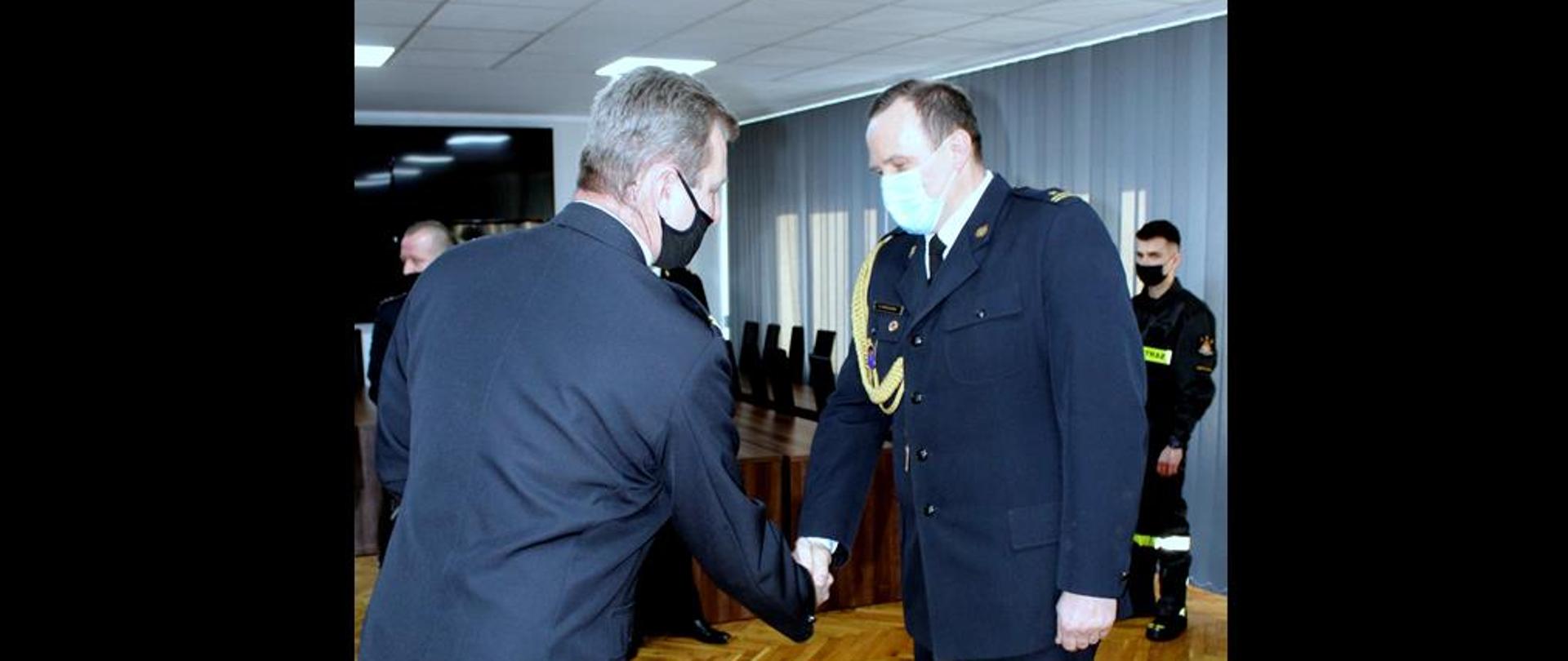 Zdjęcie przedstawia Komendanta Miejskiego Państwowej Straży Pożarnej w Częstochowie oraz Dowódcę Jednostki Ratowniczo Gaśniczej numer 4 podczas wręczenia aktu nominowania na wyższe stanowisko służbowe. Funkcjonariusze w umundurowaniu galowym podczas uścisku ręki. 