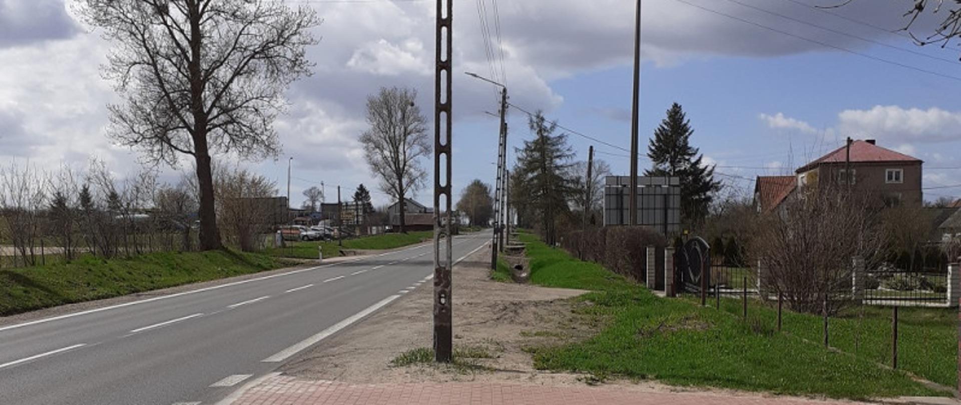 Asfaltowa droga z piaszczystym poboczem. Na wprost zjazd do posesji z czerwonego kostki betonowej 