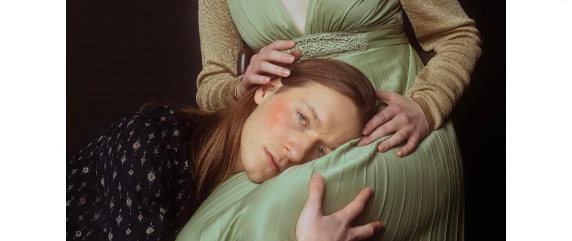 Zdjęcie z białą grubą ramką prezentuje młodego mężczyznę, który wtula się w łono kobiety. Oboje ubrani są w klasyczne stroje.