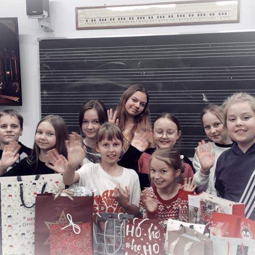 Grupa uczniów stoi na tle tablicy w pięciolinie. Na ławce przed uczniami znajdują się świąteczne torby z prezentami mikołajkowymi. Uczniowie uśmiechają się i machają rękami na powitanie.