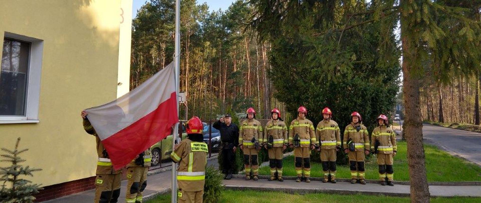 Na pierwszym planie z lewej trzej strażacy w żółtych ubraniach specjalnych i czerwonych hełmach w trakcie zawieszania flagi Państwowej na maszt. W tle w szeregu stoją pozostali strażacy, sześciu w żółtych ubraniach specjalnych i czerwonych hełmach, pierwszy z lewej w czarny, ubraniu sztabowym, jest to dowódca JRG4 w Bydgoszczy. Zbiórka przebiega na placu z kostki brukowej przed JRG4 w Bydgoszczy. W tle z prawej ulica, za strażakami drzewa. 