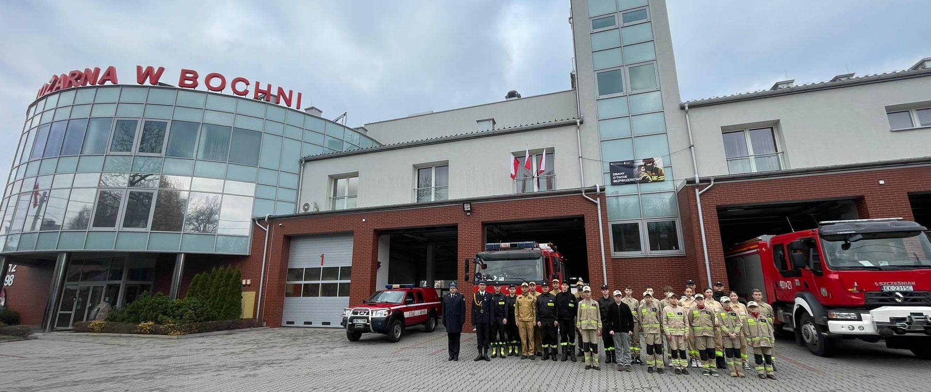 Grupa strażaków w mundurach koszarowych znajdują się przed garażami. z lewej strony szyku znajduje się dwóch strażaków w umundurowaniu wyjściowym. Pomiędzy strażakami a garażami znajduje się kilka samochodów pożarniczych. Tło stanowi budynek Komendy Powiatowej PSP w Bochni.