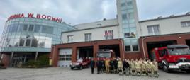 Grupa strażaków w mundurach koszarowych znajdują się przed garażami. z lewej strony szyku znajduje się dwóch strażaków w umundurowaniu wyjściowym. Pomiędzy strażakami a garażami znajduje się kilka samochodów pożarniczych. Tło stanowi budynek Komendy Powiatowej PSP w Bochni.