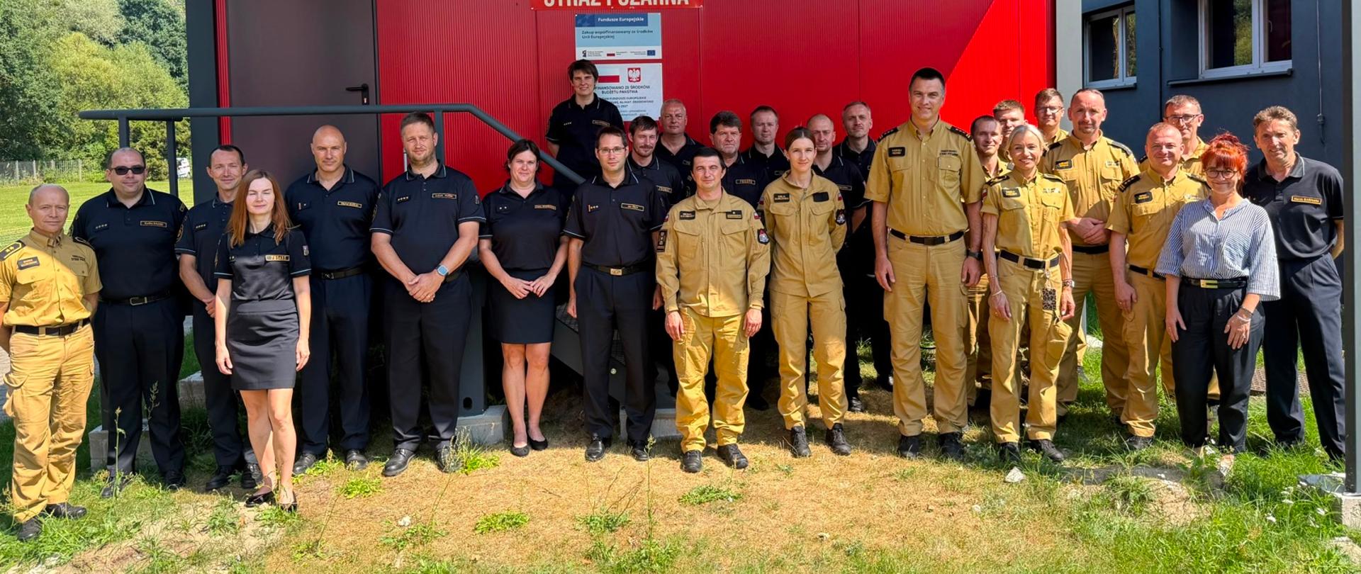 Grupa ponad dwudziestu strażaków i strażaczek w mundurach galowych oraz ćwiczebnych pozuje do wspólnego zdjęcia na tle czerwonego budynku z szyldem „PAŃSTWOWA STRAŻ POŻARNA”