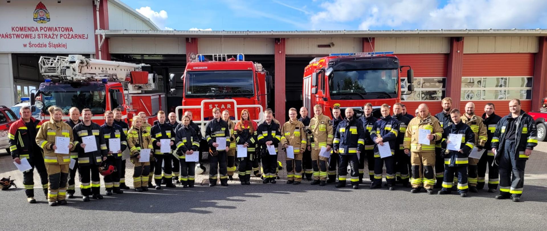 Grupa strażaków stoi na tle samochodów pożarniczych 