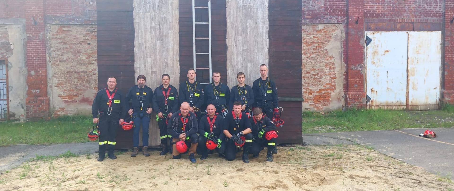 Strażacy - ratownicy Państwowej Straży Pożarnej w ubraniach koszarowych i kombinezonach z kaskami ratowniczymi i w uprzężach wykonują zadania związane ze Szkoleniem doskonalącym z ratownictwa wysokościowego realizowanego przez KSRG w zakresie podstawowym. Strażacy stoją w grupie pod wspinalnią w Komendzie Powiatowej PSP w Prudniku.