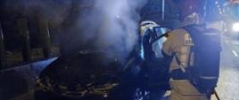 Na zdjęciu widzimy palący się samochód oraz strażaka, który gasi pożar. Zdjęcie wykonane w porze nocnej.