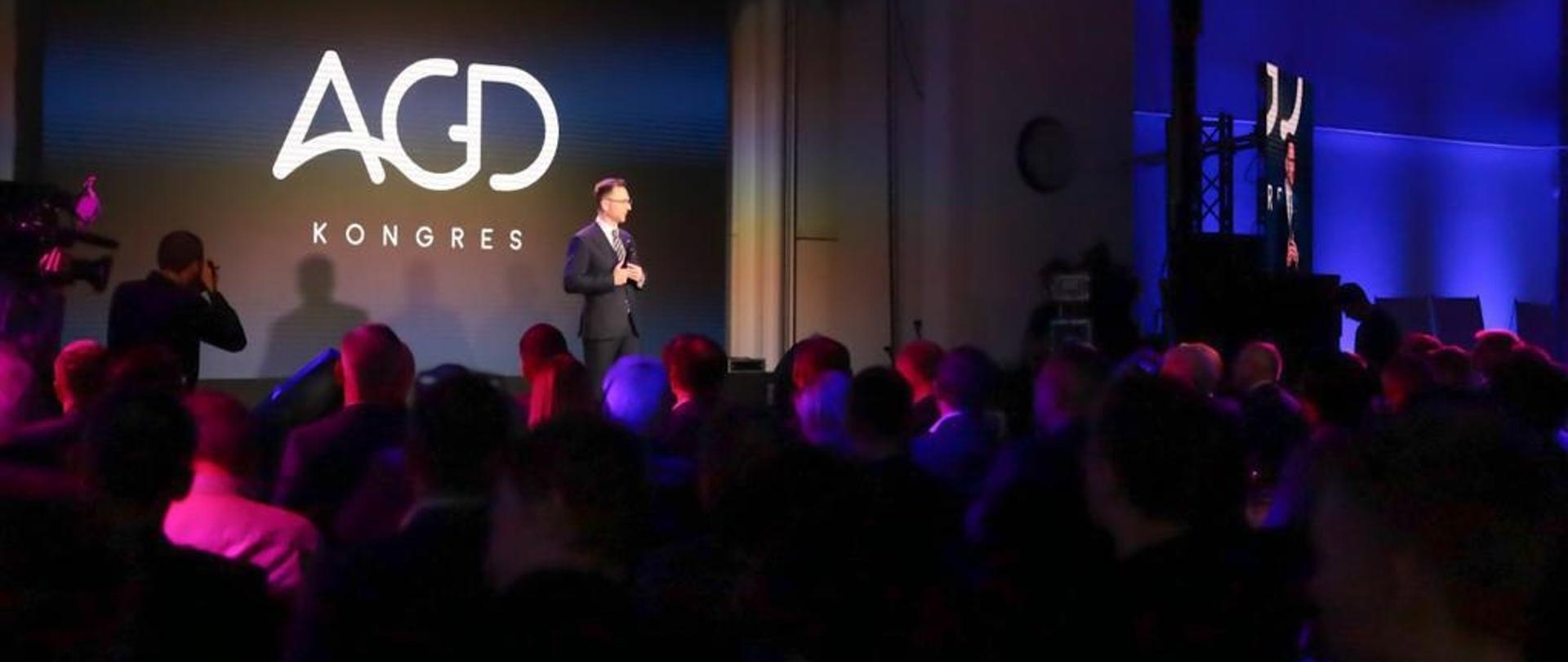 Minister rozwoju i technologii Waldemar Buda przemawiający podczas obrad Kongresu AGD, minister stoi na scenie i trzyma w dłoniach mikrofon, za jego plecami logo Kongresu