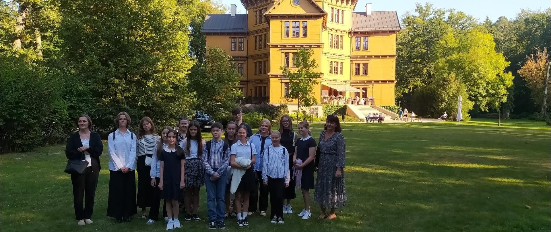 Grupowe zdjęcie uczniów i nauczycielek wykonane na tle drewnianego pałacu myśliwskiego Radziwiłłów w Antoninie w kolorze żółtym, otoczonego zielenią. Osoby pozują na trawniku w słoneczny dzień podczas wycieczki szkolnej. Uczniowie ubrani są w eleganckie stroje. 