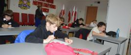 Świetlica komendy. Przy stolikach siedzą uczestnicy eliminacji, którzy rozwiązuja test pisemny. W tle ściana z nazwą komendy oraz stojak z biało-czerwonymi flagami.