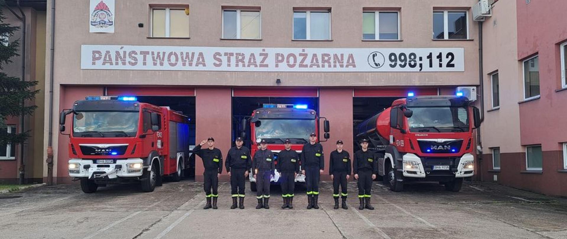 Na tle remizy i bojowego samochodu strażackiego stoi w rzędzie kilki strażaków. Jeden z nich salutuje. 