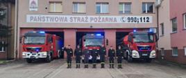Na tle remizy i bojowego samochodu strażackiego stoi w rzędzie kilki strażaków. Jeden z nich salutuje. 