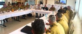 W sali przy stołach siedzą uczestnicy odprawy - funkcjonariusze PSP w mundurach służbowych koloru musztardowego
