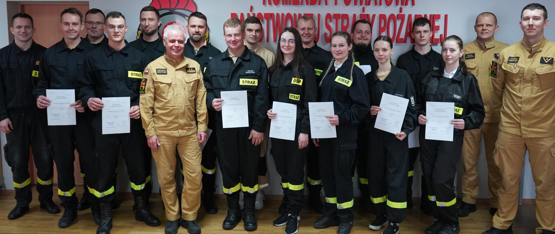 Na tle ściany, na której widnieje czerwony napis "Komenda Powiatowa Państwowej Straży Pożarnej w Rawiczu" i logo PSP, w dwuszeregu stoją strażacy uczestniczący w kursie kwalifikowanej pierwszej pomocy. Wsród nich są 4 kobiety. Wspólnie z zastępcą komendanta i członkami komisji egzaminacyjnej pozuja do pamiątkowego zdjęcia. 