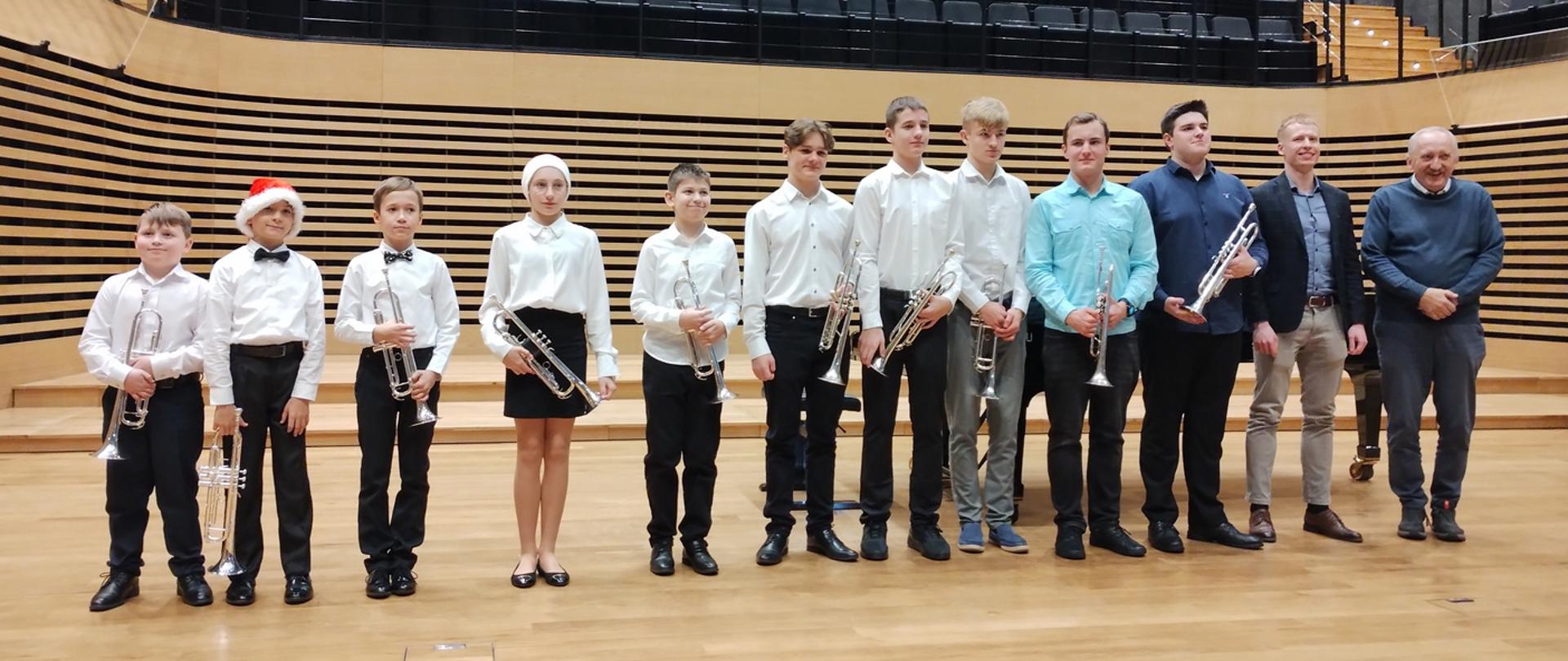 Czterech chłopców, jedna dziewczynka i pięciu nastolatków oraz dwóch mężczyzn stoi w jednym rzędzie na estradzie sali koncertowej. Wszyscy uczniowie trzymają przed sobą trąbki. Dziewczynka i jeden chłopiec mają na głowach czapki Mikołaja.