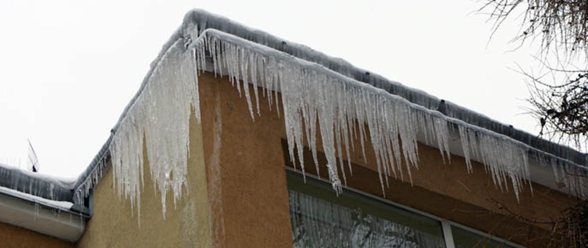 Część elewacji budynku, na górze którego widoczne są zwisające sople. 