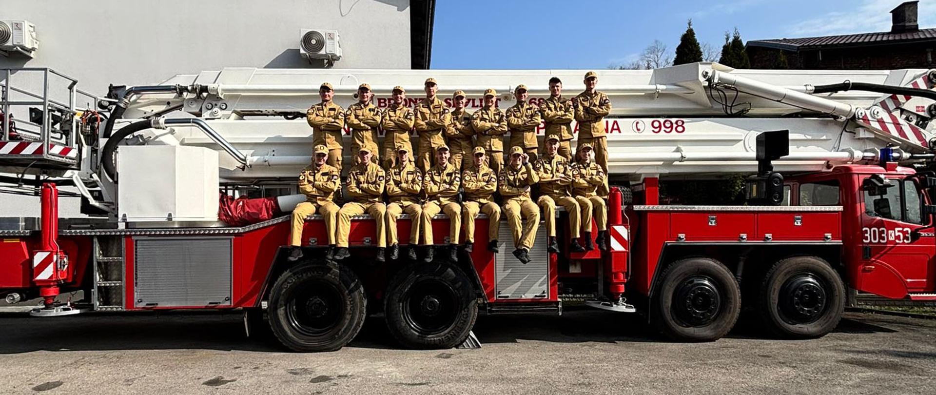 Grupa kadetów Centralnej Szkoły Państwowej Straży Pożarnej siedzi i stoi na dużym pojeździe pożarniczym podczas wspólnego, pamiątkowego zdjęcia. Strażacy ubrani są w jednolite mundury koloru piaskowego