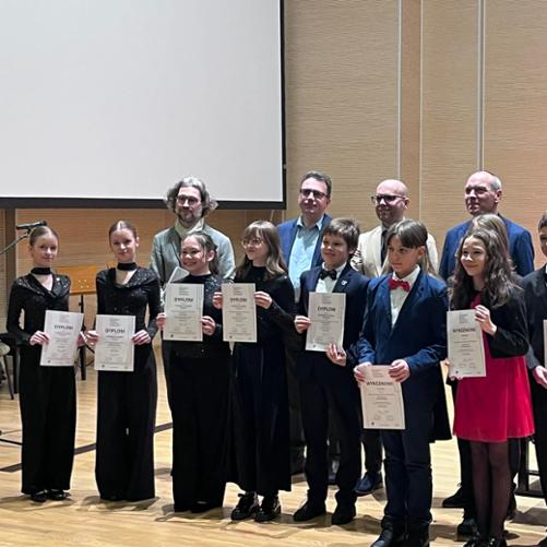 Zdjęcie grupowe laureatów XVII Ogólnopolskiego Konkursu Kameralistyki Fortepianowej w Toruniu. W rzędzie dalej stoją mężczyźni - jurorzy. Z przodu stoi 10 pianistów obu płci. Większość ubrana jest na czarno, wszyscy odświętnie. Jedna dziewczynka ma czerwoną spódnicę. Młodzież trzyma w ręku dyplomy A4.