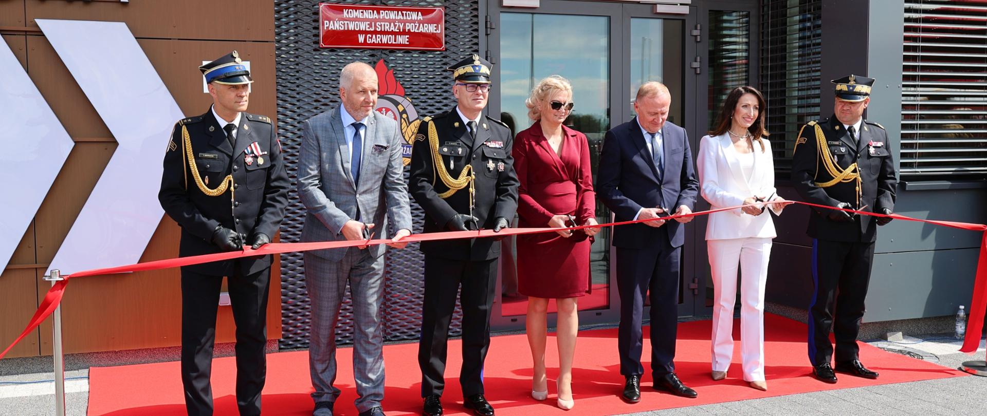 Uroczyste przecięcie czerwonej wstęgi przed głównym wejściem do nowego budynku Komendy Powiatowej Państwowej Straży Pożarnej w Garwolinie. W ceremonii biorą udział m.in. Sekretarz Stanu Czesław Mroczek, Starosta Iwona Kurowska oraz wysocy rangą oficerowie PSP w mundurach galowych.