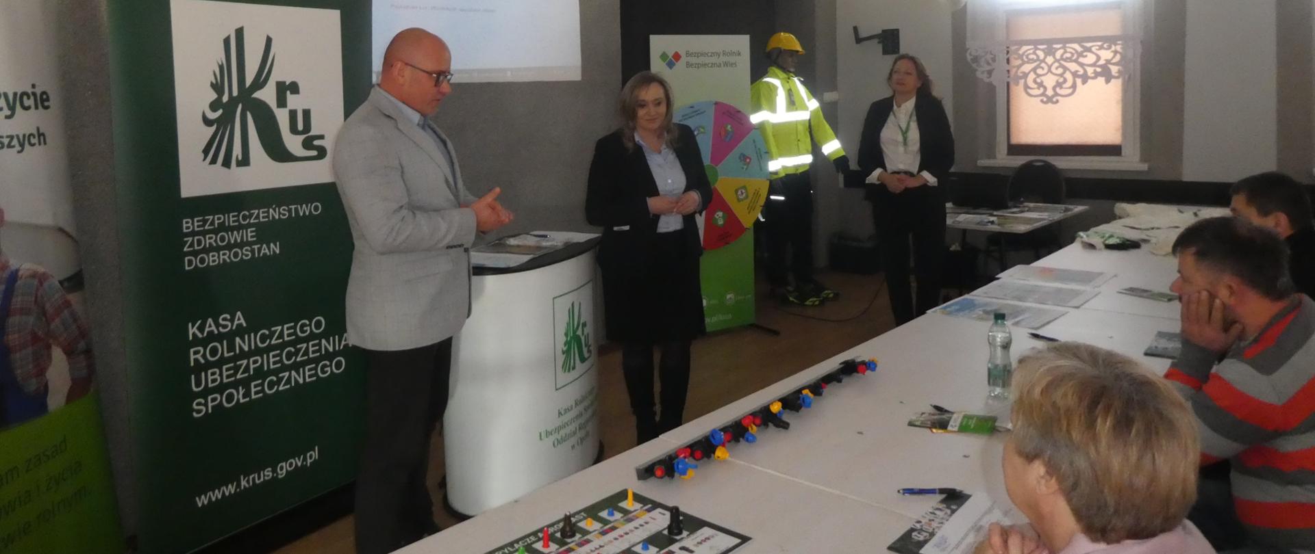 Mężczyzna i dwie kobiety stoją na tle tablic promujących KRUS i ekranu, pomiędzy nimi stoi manekin w stroju ochronnym z odblaskami. Przed nimi siedzi grupa osób przy połączonych ławkach i słuch przemawiającego mężczyzny.