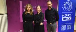 Zdjęcie pozowane. Od lewej: Aniela Giecko, Aleksandra Makiewicz, Tomasz Banaszkiewicz stoją na tle banerów. Obok nauczyciela niebieski baner z białą grafiką i napisami. Rysunek twarzy kobiety. Z prawej strony jest ona otoczona gałęzią z liśćmi, z lewej są to gwiazdki. Niżej napis "NAUKA UMCS", następnie nauka@mail.umcs.pl, niżej dwa kody QR., niżej logo UMCS i logo ATHENA.