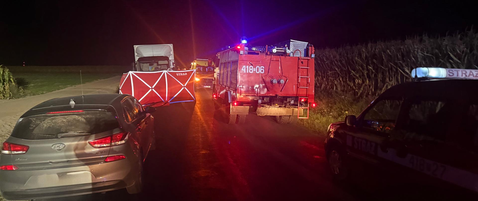 Pojazdy osobowe na jezdni wraz z pojazdem straży pożarnej na tle parawanu i pogotowia ratunkowego i straży pożarnej.