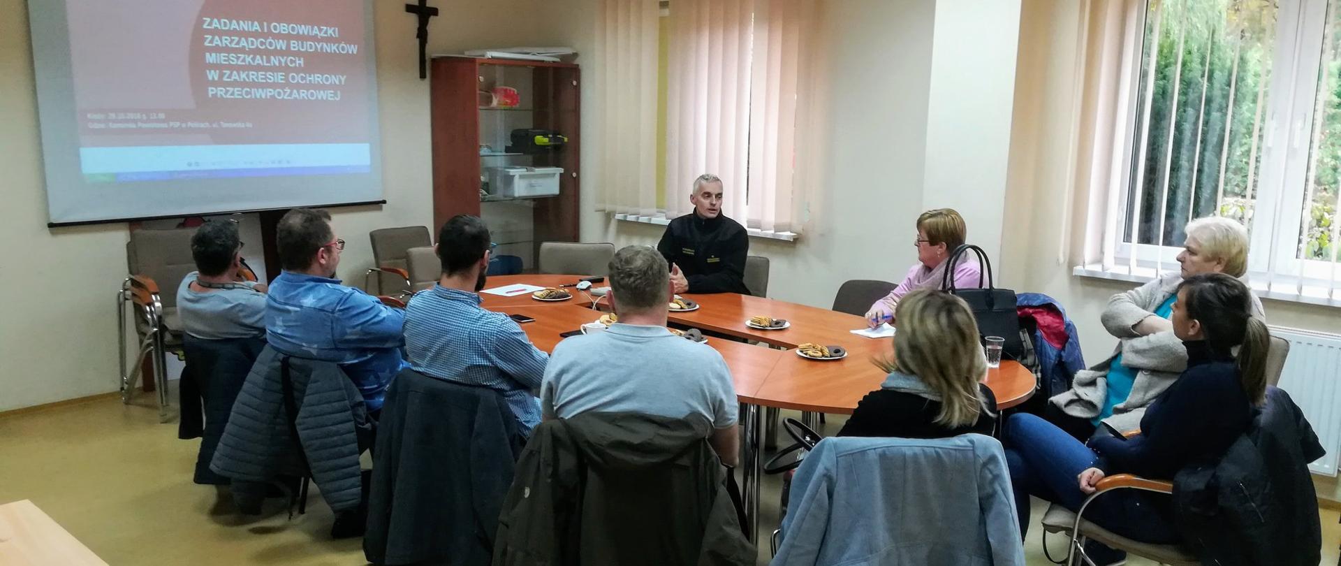 Dziewięciu ludzi przy owalnym stole, na ścianie ekran z prazentacją