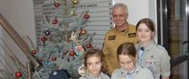 Na korytarzu komendy powiatowej PSP w Rawiczu do wspólnego zdjęcia pozują zuchy wraz z zastępcą komendanta powiatowego PSP. Wszyscy mają na sobie mundury. Chłopiec - zuch na głowie ma czapke dżokejkę. W rękach trzyma lampion, z Betlejemskim Światłem Pokoju. Dziewczynka w okularach, stojąca obok, ma w rękach tabliczkę z życzeniami. Z lewej strony stoi ubrana w ozdoby, choinka.