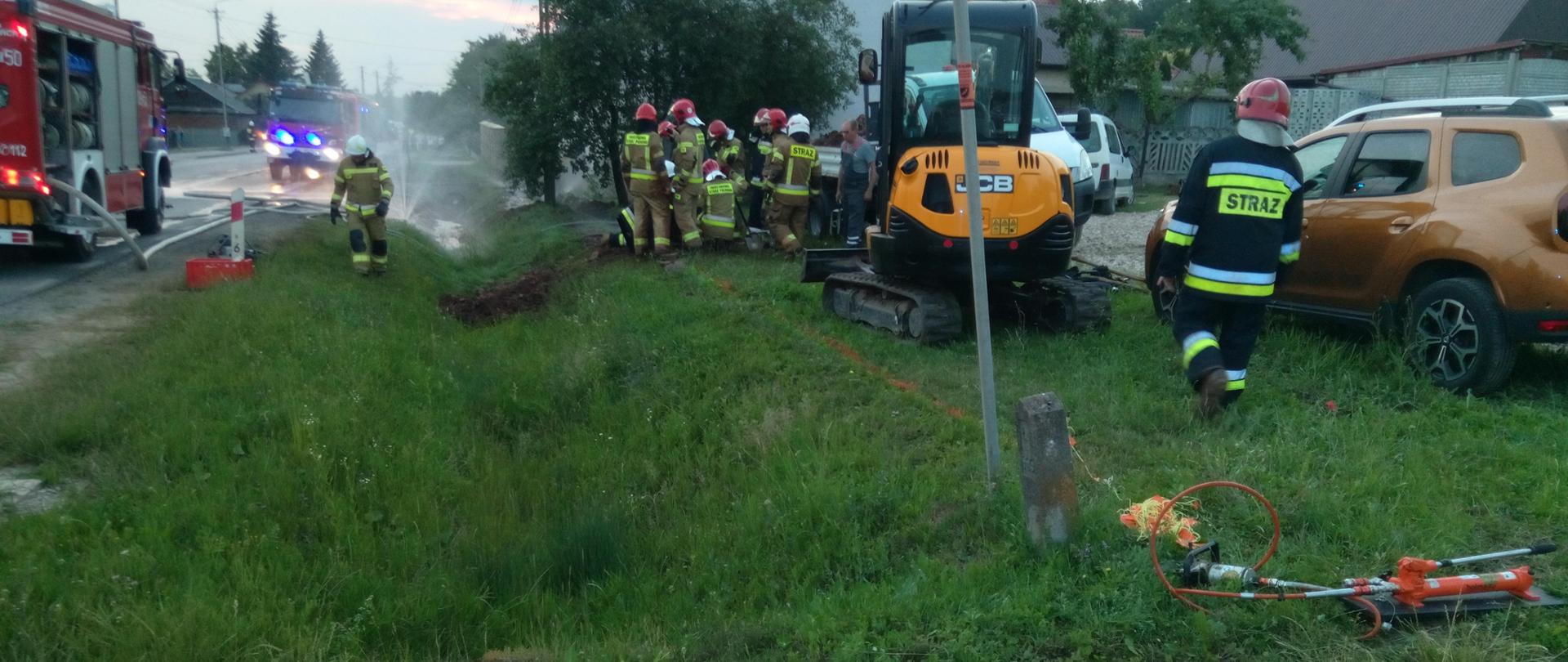 Na zdjęciu widać drogę na której stoją samochody strażackie. Na terenie przyległym do drogi stoi koparka oraz grupa strażaków pracująca przy uszczelnieniu rurociągu.